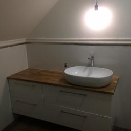 USŁUGI BUDOWLANO-REMONTOWE,LASKOWSKI PIOTR - Nowoczesna umywalka nablatowa w owalnym kształcie, osadzona na drewnianym blacie szafki z białymi frontami w łazience na poddaszu, oświetlona pojedynczą lampą wiszącą.
