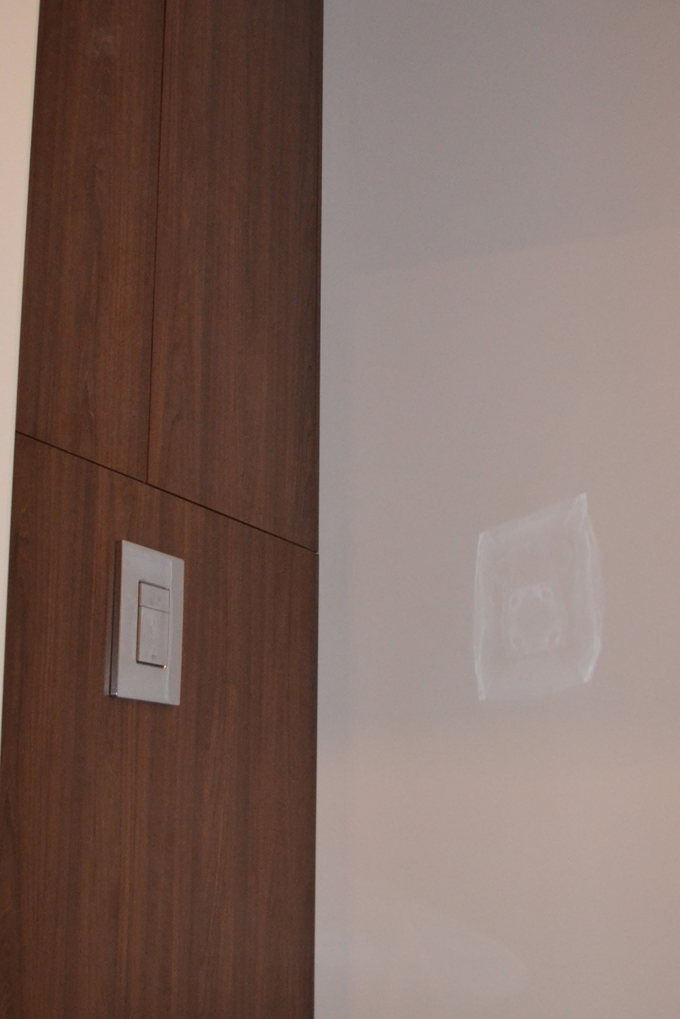Ciemnobrązowe, drewniane drzwi z zamontowanym, kwadratowym włącznikiem światła na ścianie. Widoczny ślad po tapecie.