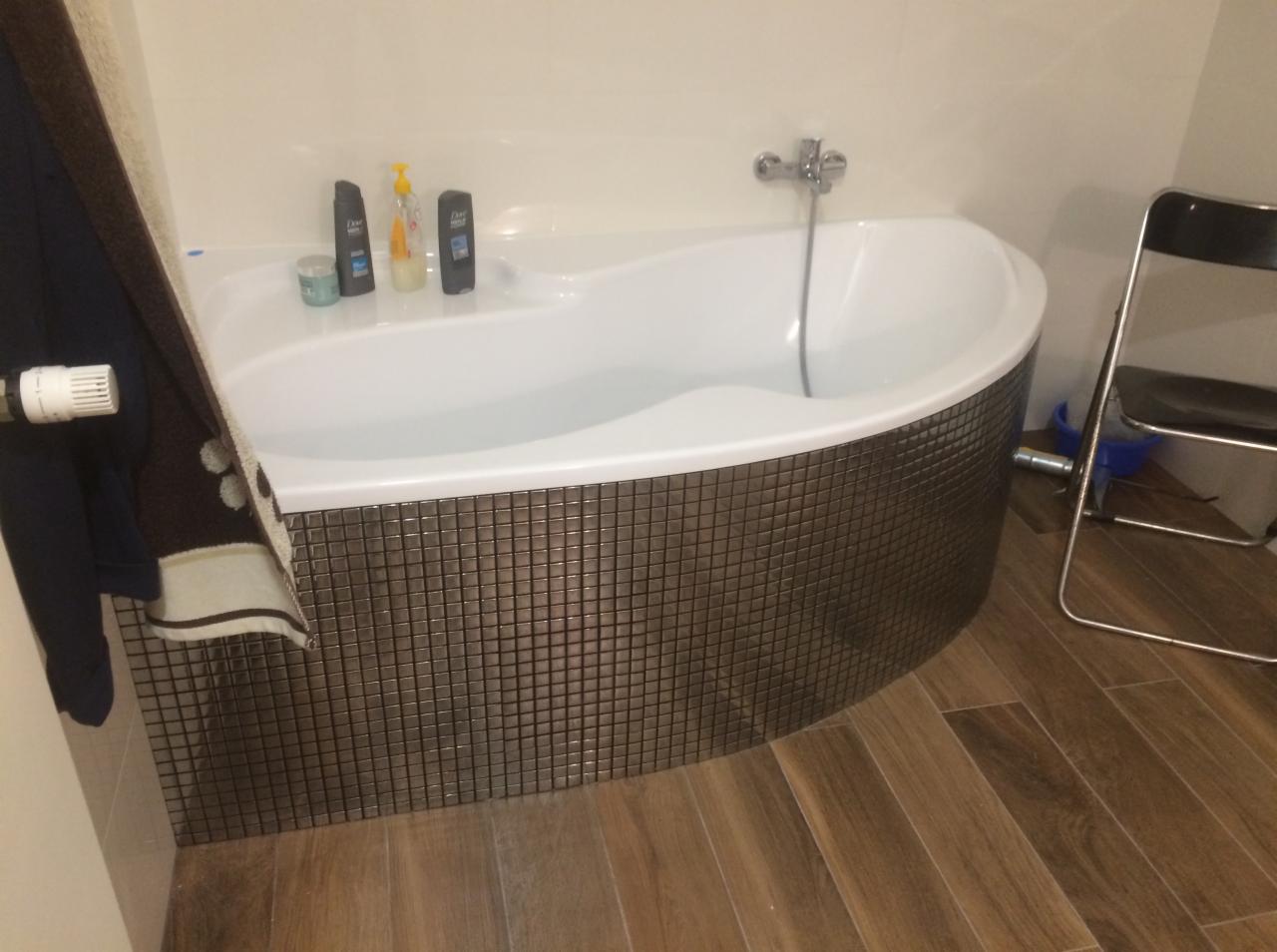 Biała wanna obudowana mozaiką w odcieniach brązu, z widocznymi kosmetykami na krawędzi i drewnopodobną podłogą, sfotografowana z niskiego kąta.