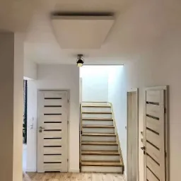 Płaski, biały panel grzewczy zamontowany na suficie w nowoczesnym przedpokoju z drewnianymi schodami i dwiema parami drzwi.