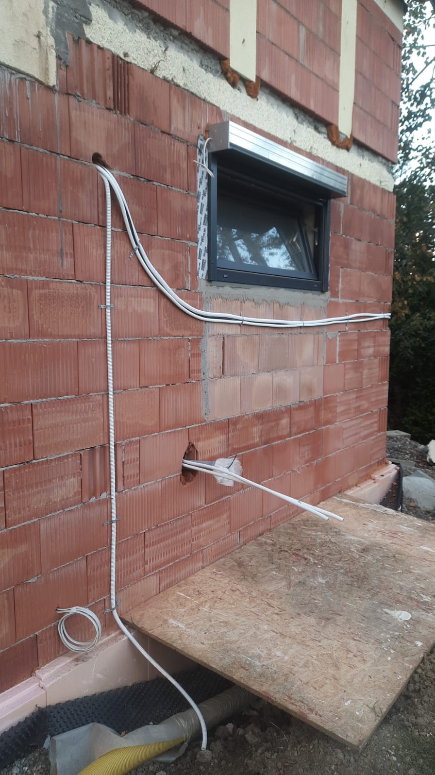 Czerwona ściana z pustaków z widocznymi przewodami elektrycznymi, rurami i oknem z roletą zewnętrzną w trakcie instalacji.