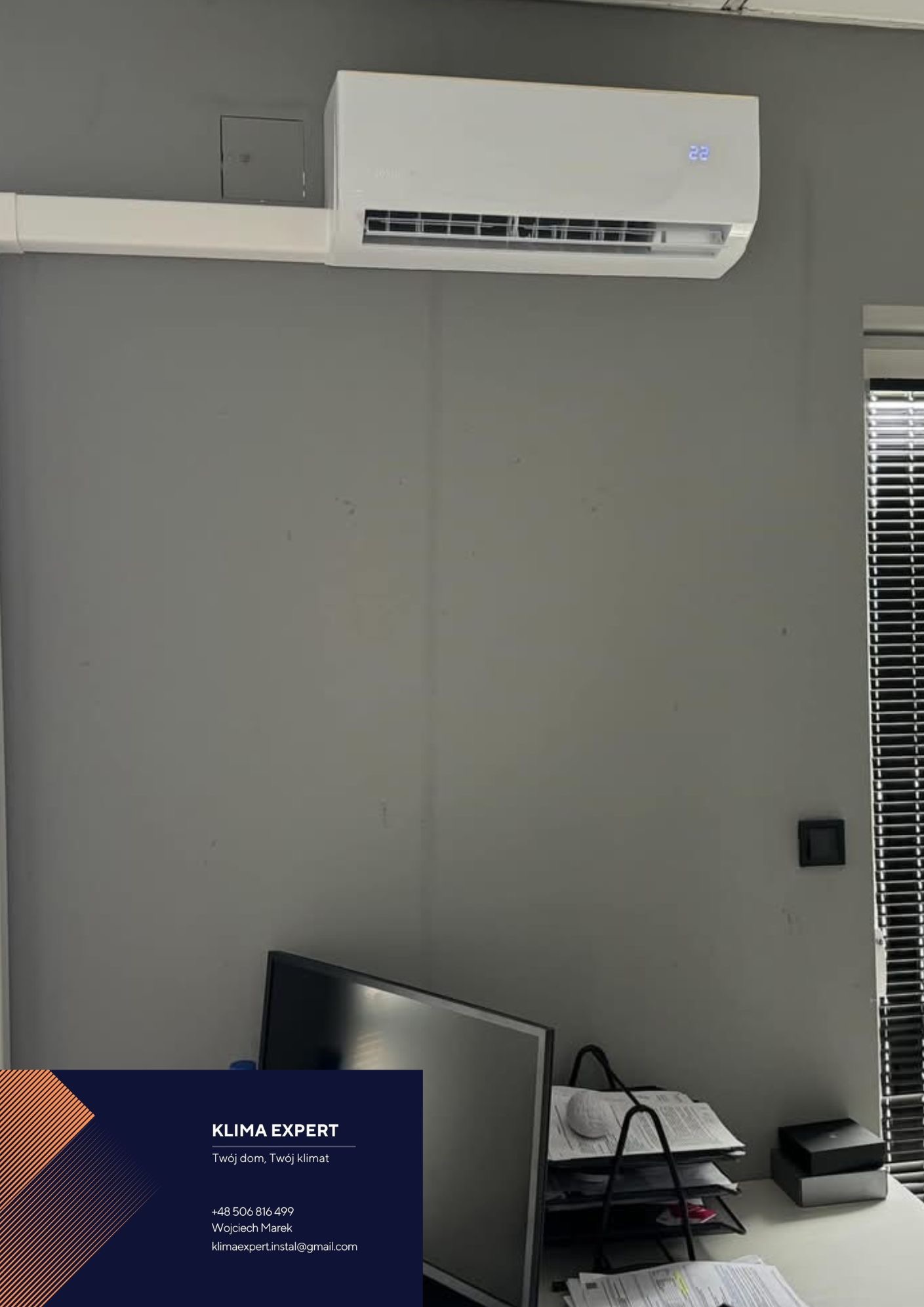 Biały klimatyzator ścienny z wyświetlaczem temperatury (22 stopnie), zamontowany nad biurkiem z monitorem i wizytówką firmy Klima Expert na szarej ścianie.