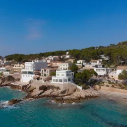 Widok z lotu ptaka na nadmorskie domy z białymi elewacjami, położone na klifowym wybrzeżu z turkusową wodą i piaszczystą plażą, otoczone zielenią drzew i wzgórz.