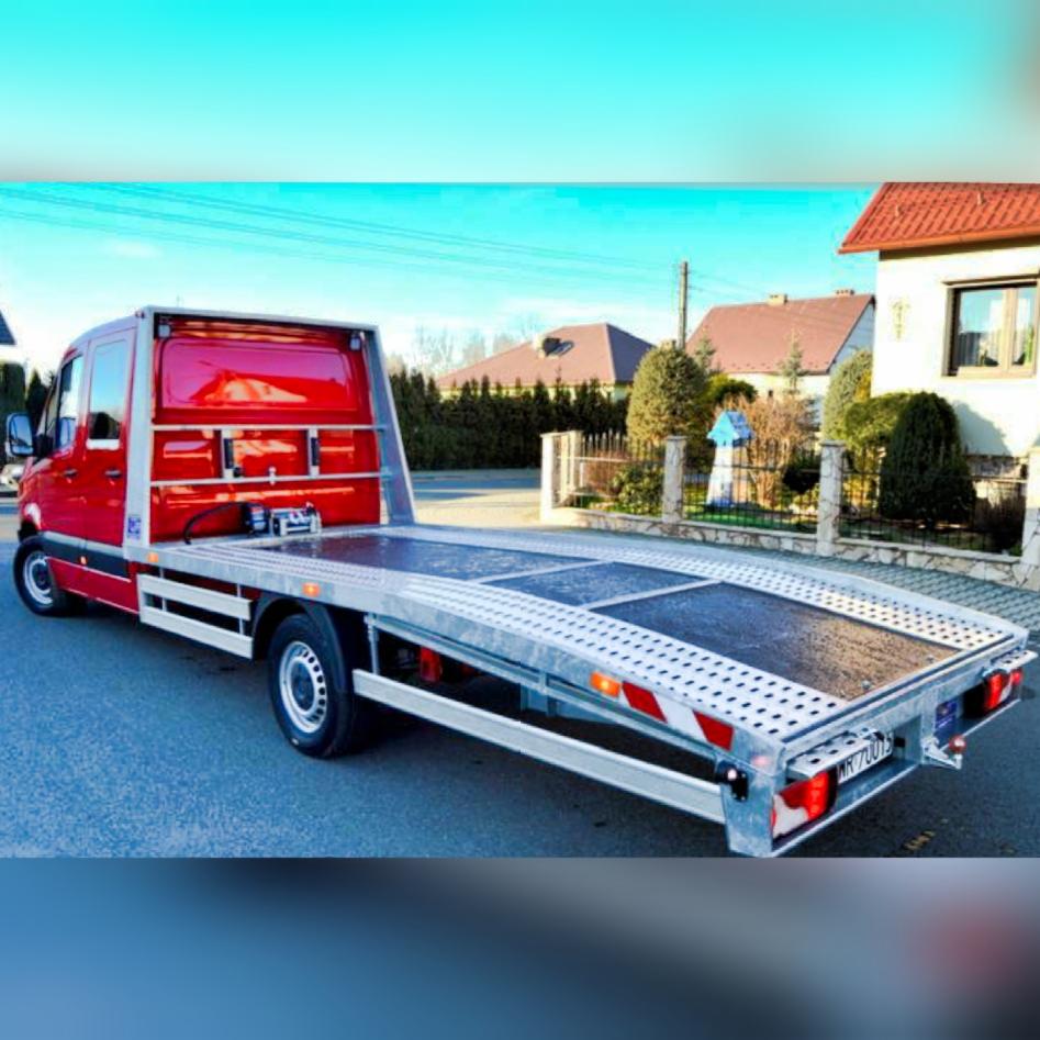 Czerwona laweta na ulicy, widok z tyłu, w tle domy jednorodzinne, błękitne niebo. Platforma lawety pusta, gotowa do załadunku pojazdu.