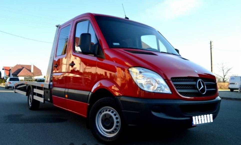 Czerwona laweta Mercedes-Benz Sprinter z platformą, zaparkowana na ulicy. Widoczne budynki w tle i światła odbijające się na karoserii.