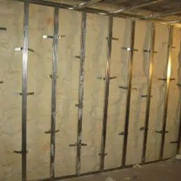 Ściana wewnętrzna z metalowym stelażem wypełniona jasnobeżową pianką izolacyjną PUR, widoczne metalowe łączniki stelaża.