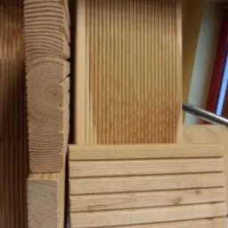 Ułożone deski tarasowe z widocznym przekrojem słojów drewna, ukazujące fakturę i naturalny odcień jasnego drewna. Deski ułożone pionowo i poziomo.