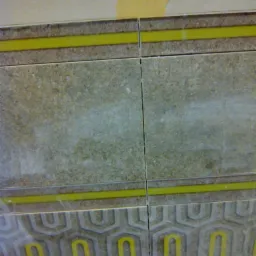 Fragment ściany z ułożonymi płytkami w odcieniach szarości i beżu, z żółtymi akcentami dekoracyjnymi i fragmentem oderwanej tapety u góry.