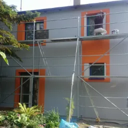 Praca na rusztowaniu przy szarej elewacji budynku z pomarańczowymi obramowaniami okien. Malarz w oknie, widoczne rośliny w ogrodzie. Dom z pomalowaną elewacją.