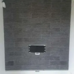 Wykończona ściana z szarej, poziomej okładziny kamiennej z otworem na TV. Białe ściany boczne i sufit. Podłoga w trakcie prac wykończeniowych.