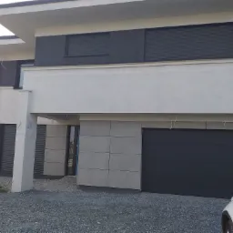 Nowoczesny dom jednorodzinny z szarą elewacją, grafitową bramą garażową i żaluzjami zewnętrznymi, wykończony drobnym żwirem.