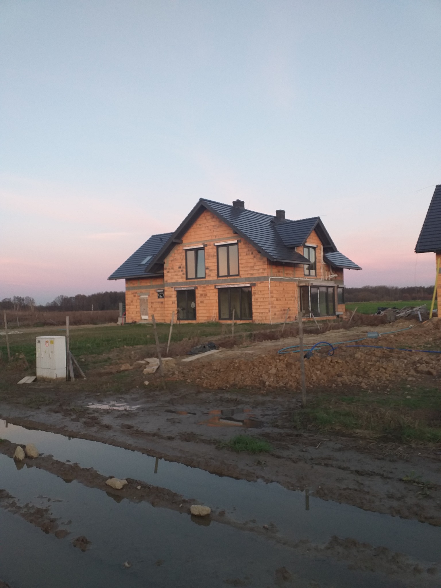 Surowy stan budowy domu murowanego z widoczną konstrukcją ścian z cegły, czarnym dachem i oknami bez wykończenia, na tle zachodzącego słońca i błotnistego terenu.