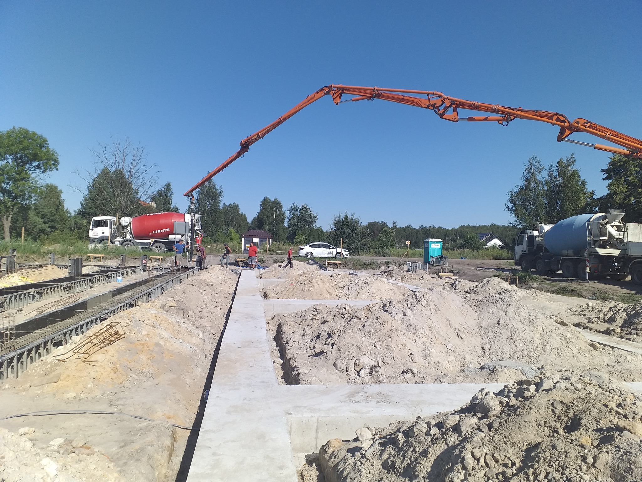 Wylewanie fundamentów domu murowanego z użyciem pompy do betonu; widoczne szalunki, zbrojenia, ekipa budowlana i betonomieszarki w tle.