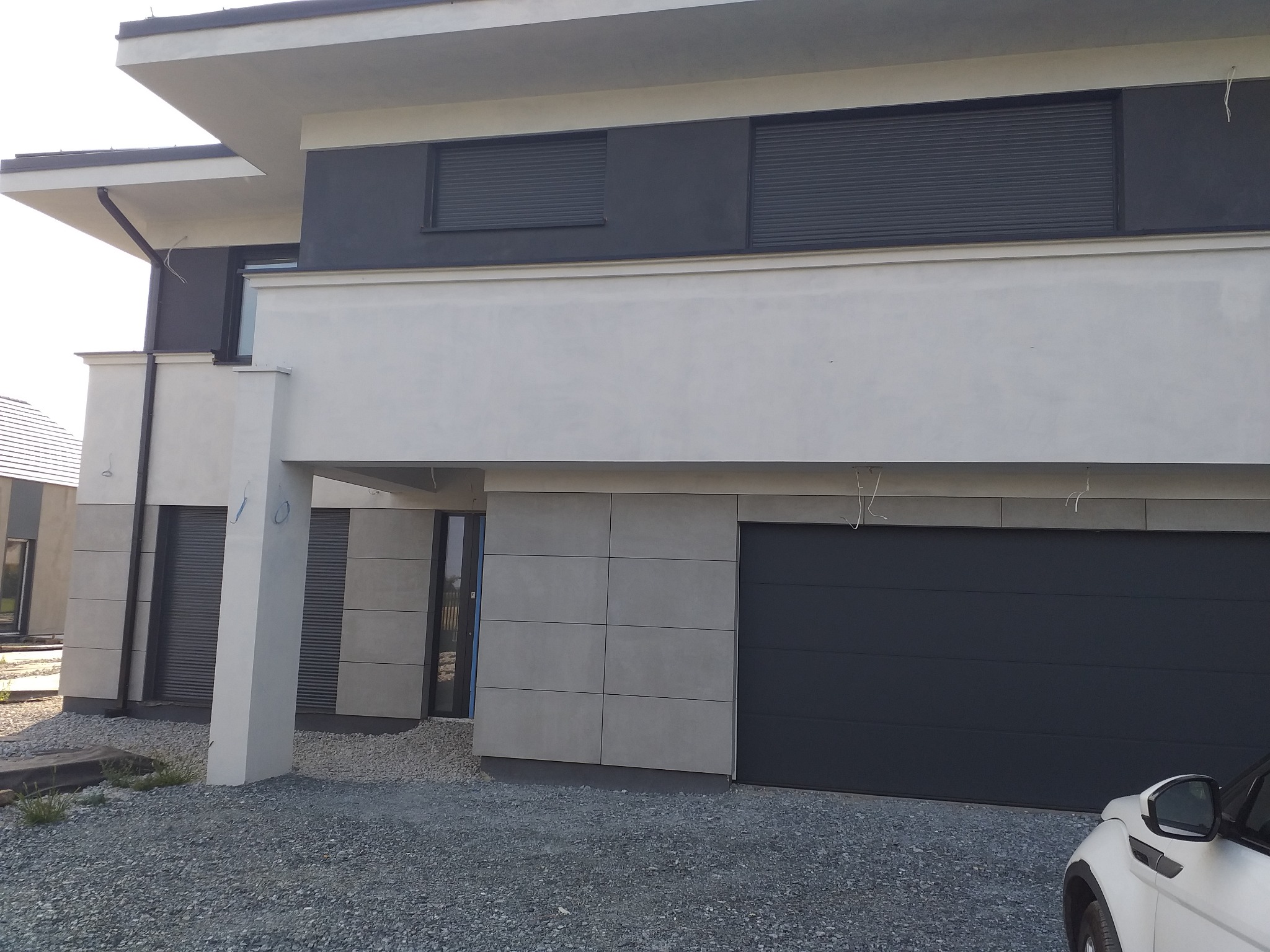 Nowoczesny dom jednorodzinny z szarą elewacją, grafitową bramą garażową i żaluzjami zewnętrznymi, wykończony drobnym żwirem.