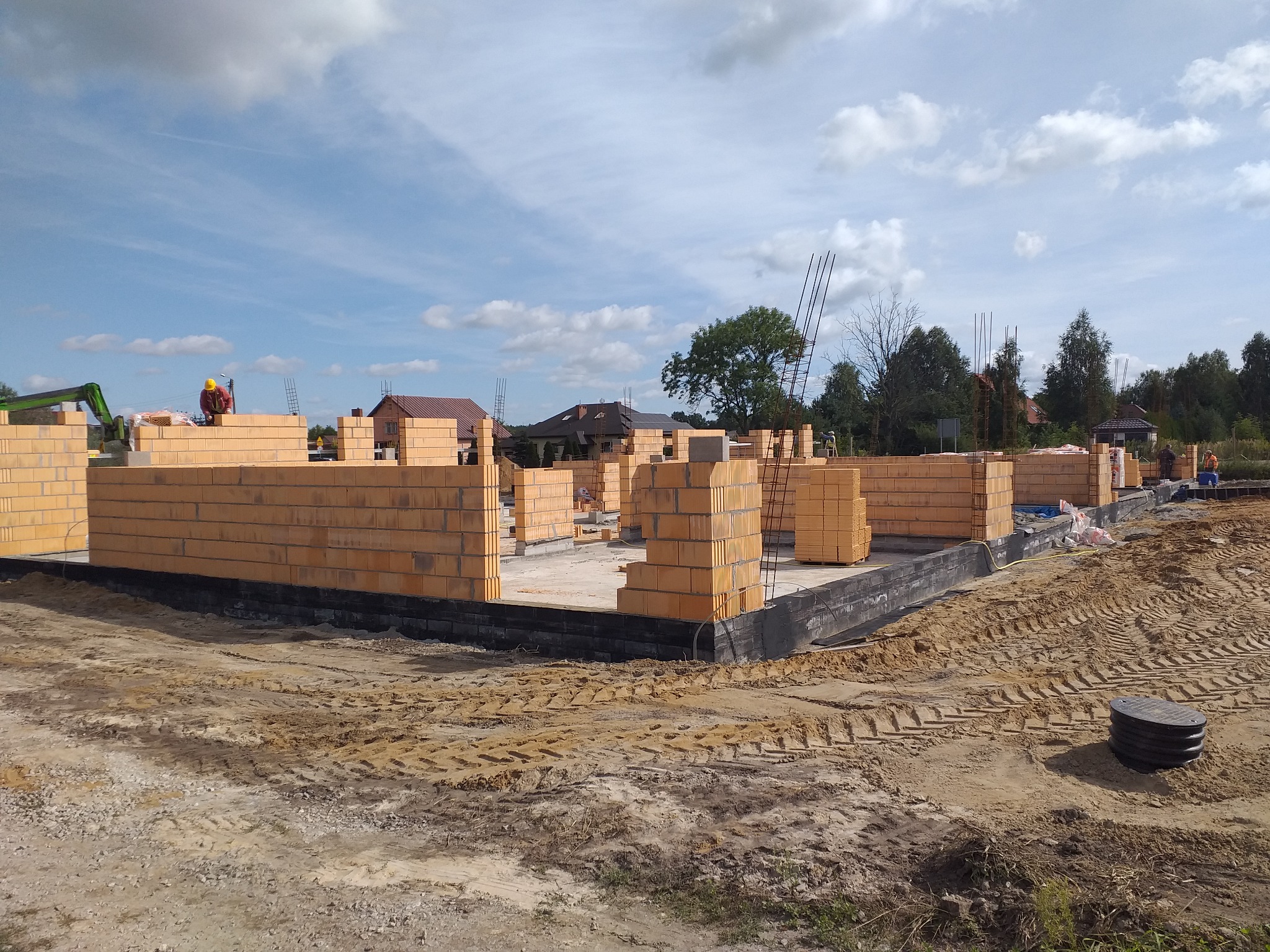 Wznoszenie ścian parteru z bloczków na fundamencie domu jednorodzinnego, widoczne zbrojenia i pracownik w kasku na budowie.