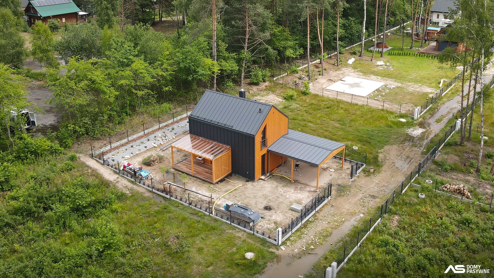 Nowoczesny dom szkieletowy z czarną elewacją i drewnianymi akcentami, widok z lotu ptaka, otoczony zielenią i ogrodzeniem. Widoczny taras z zadaszeniem.