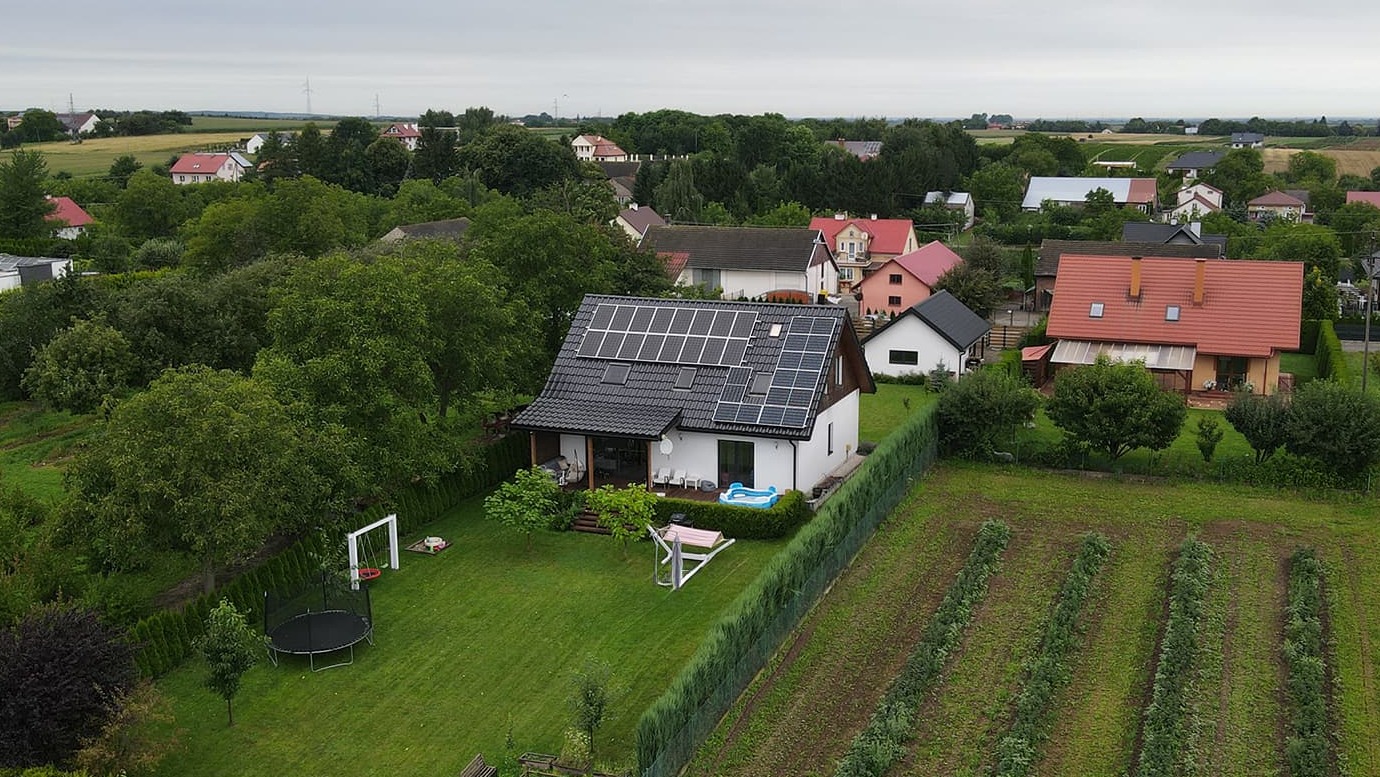 Nowoczesny dom z panelami słonecznymi na dachu, widok z lotu ptaka. W tle zabudowa wiejska i pola uprawne. Na pierwszym planie zadbany ogród z trampoliną.