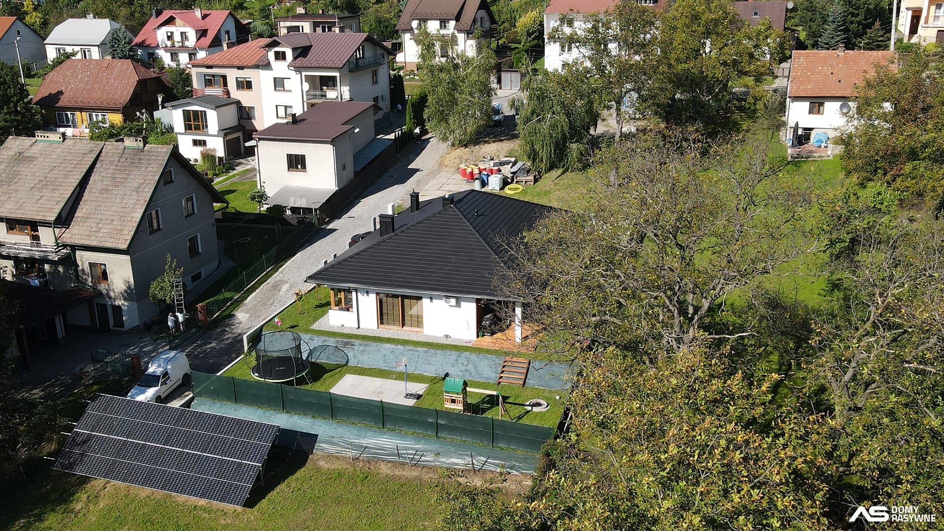 Nowoczesny dom z czarnym dachem i panelami słonecznymi, otoczony zielonym ogrodem z trampoliną i placem zabaw, widok z góry na tle zabudowy mieszkalnej.