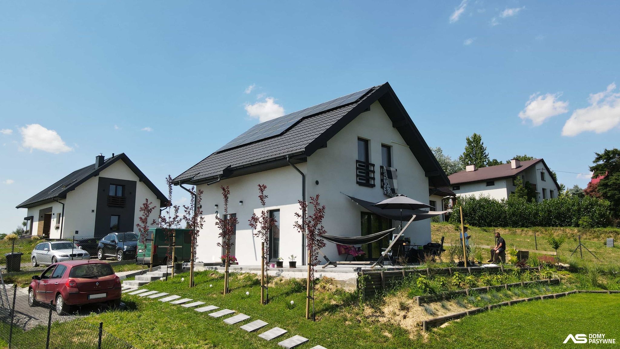 Biały dom z panelami słonecznymi na dachu, widok z ogrodu z elementami architektury krajobrazu i sąsiednimi budynkami w tle, słoneczny dzień.