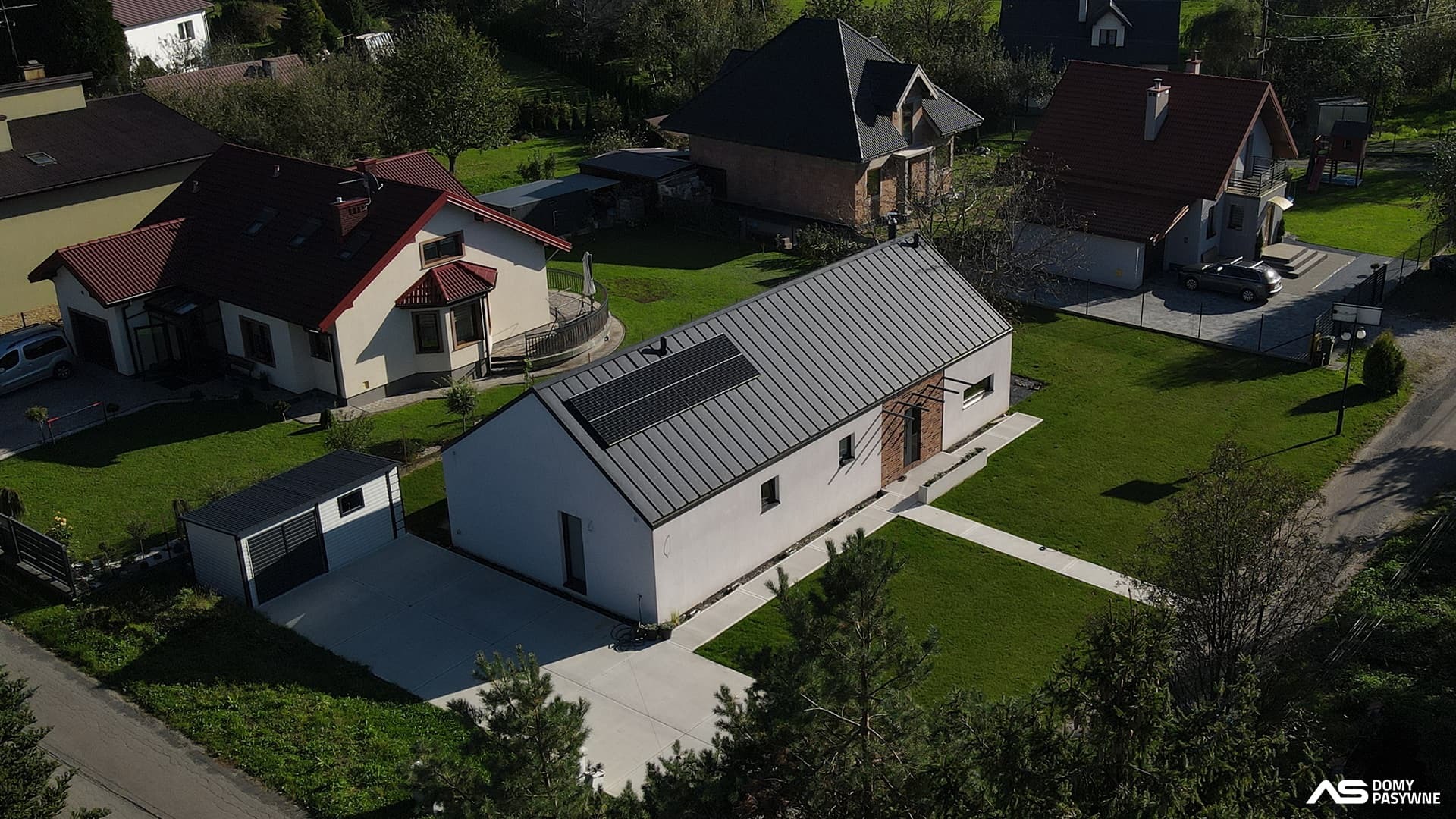 Nowoczesny dom pasywny z panelami słonecznymi na dachu, widziany z lotu ptaka. Biała elewacja, szara blacha, minimalistyczny design, otoczony zielonym trawnikiem i innymi domami.