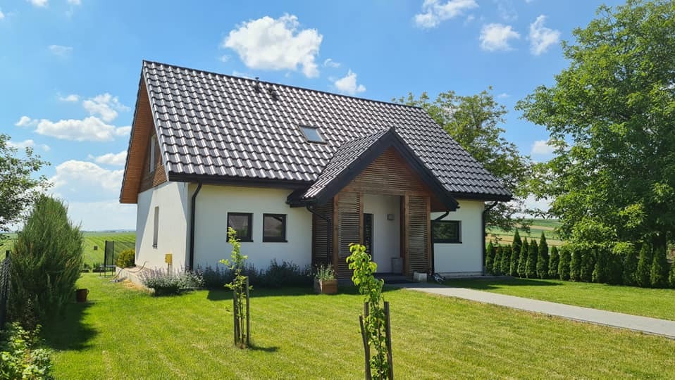 Gotowy dom szkieletowy z czarną dachówką i drewnianym wykończeniem wejścia, otoczony zielonym trawnikiem i drzewami w słoneczny dzień. Widok na pola w tle.