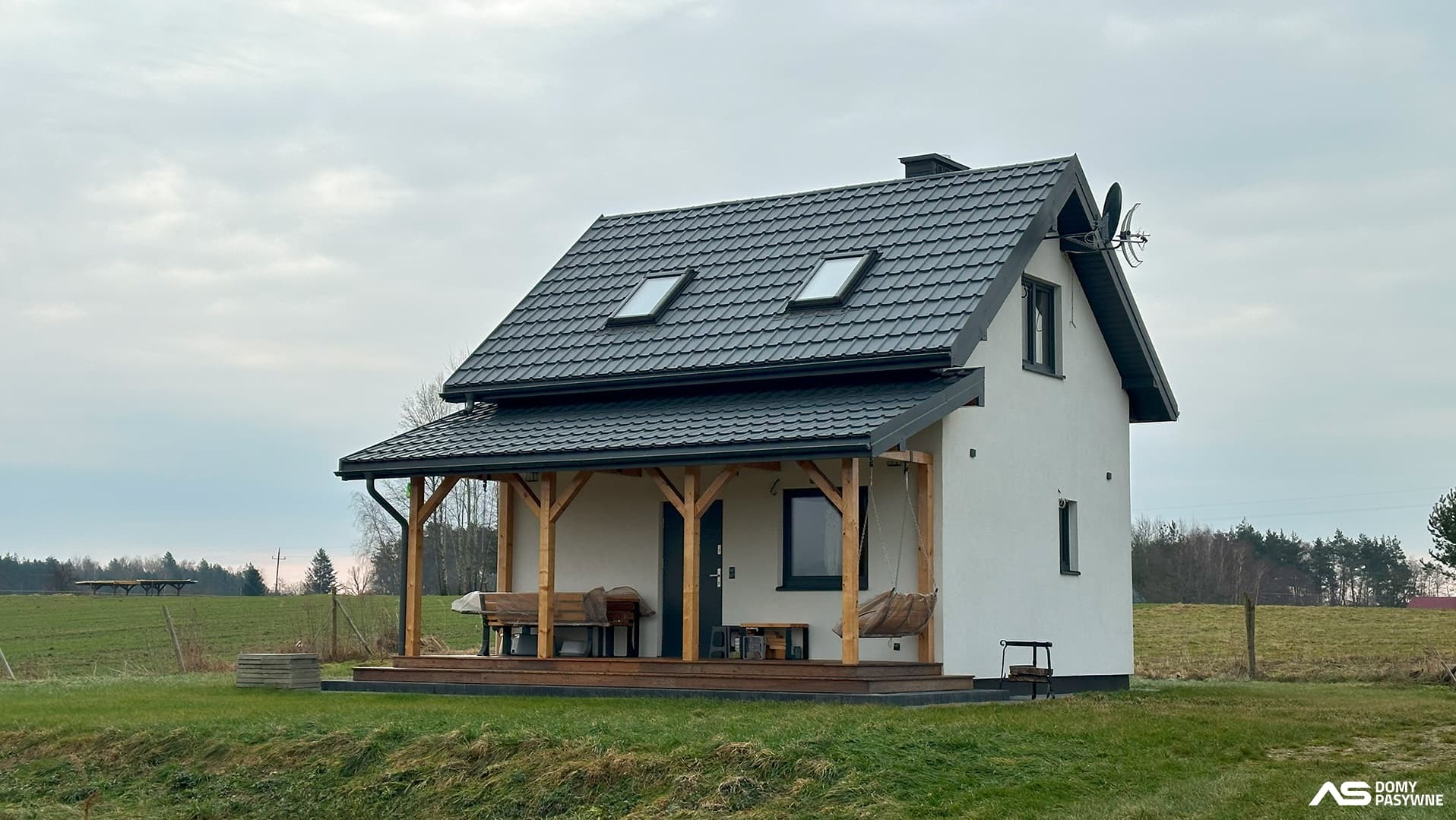 Gotowy dom szkieletowy z drewnianym tarasem i huśtawką, na tle zielonej łąki. Dach pokryty czarną blachodachówką z oknami połaciowymi. Widoczne logo firmy.