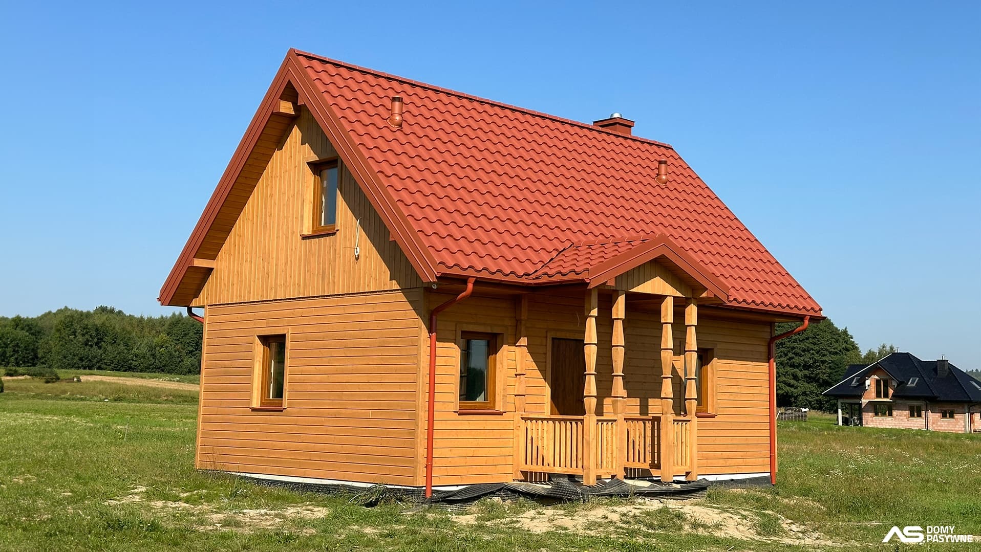 Gotowy dom szkieletowy z drewnianą elewacją i czerwonym dachem na tle zielonej łąki i błękitnego nieba. Widoczny fragment tarasu z kolumnami.