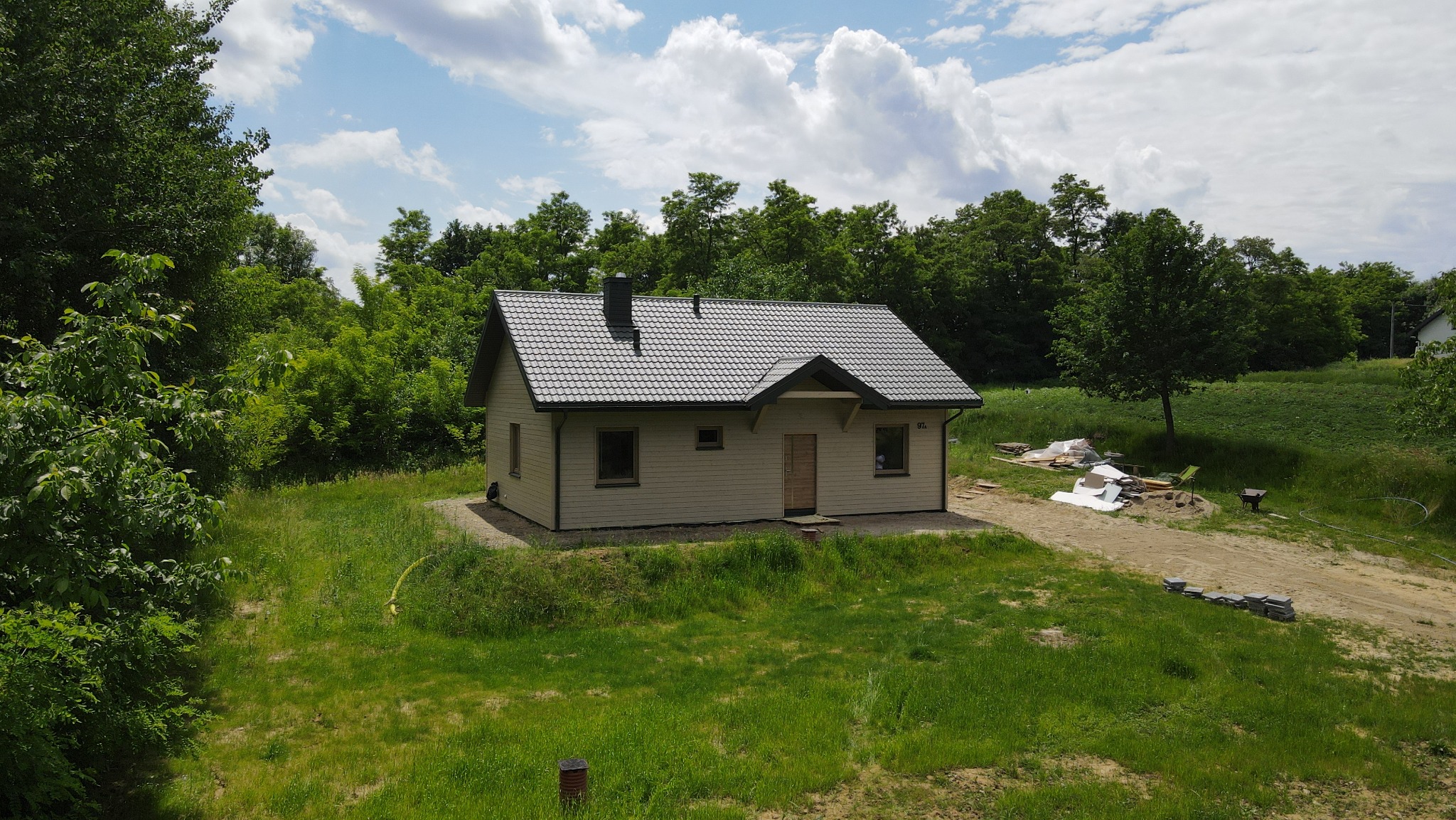 Gotowy, parterowy dom szkieletowy z ciemną dachówką, otoczony zielenią. Widoczne materiały budowlane obok domu. Ujęcie z góry, w słoneczny dzień.
