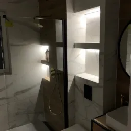 Nowoczesna łazienka z białymi płytkami imitującymi marmur, kabiną prysznicową typu walk-in z czarną ramą, złotą armaturą, podświetlanymi wnękami w ścianie i toaletą w zabudowie.