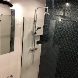 Nowoczesna kabina prysznicowa ze szklaną ścianką, chromowaną armaturą i czarną płytką z geometrycznym wzorem, oświetlona punktowym światłem.