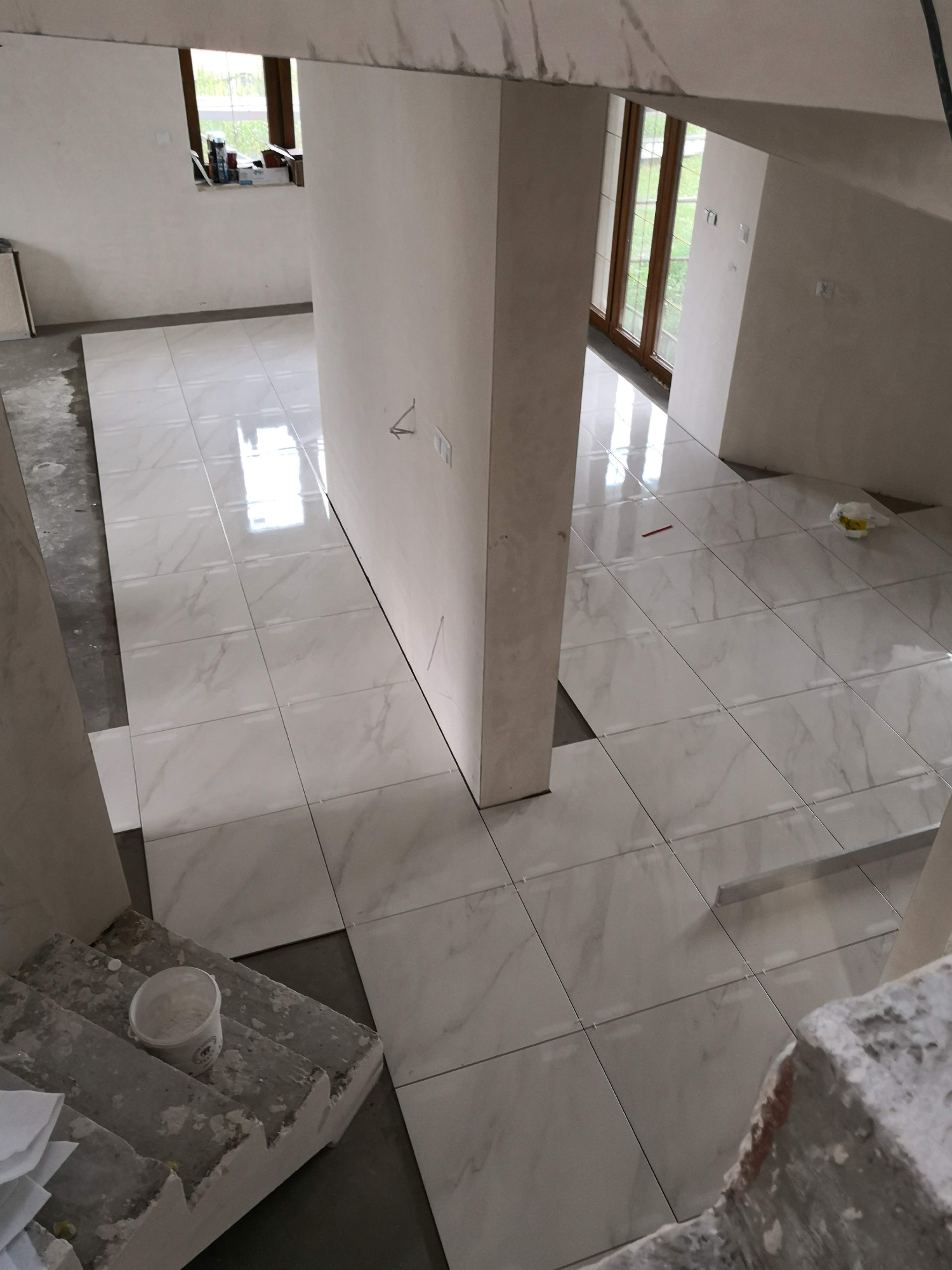 Układanie białych płytek imitujących marmur na podłodze w trakcie remontu, widoczny fragment schodów i wiadro z zaprawą.