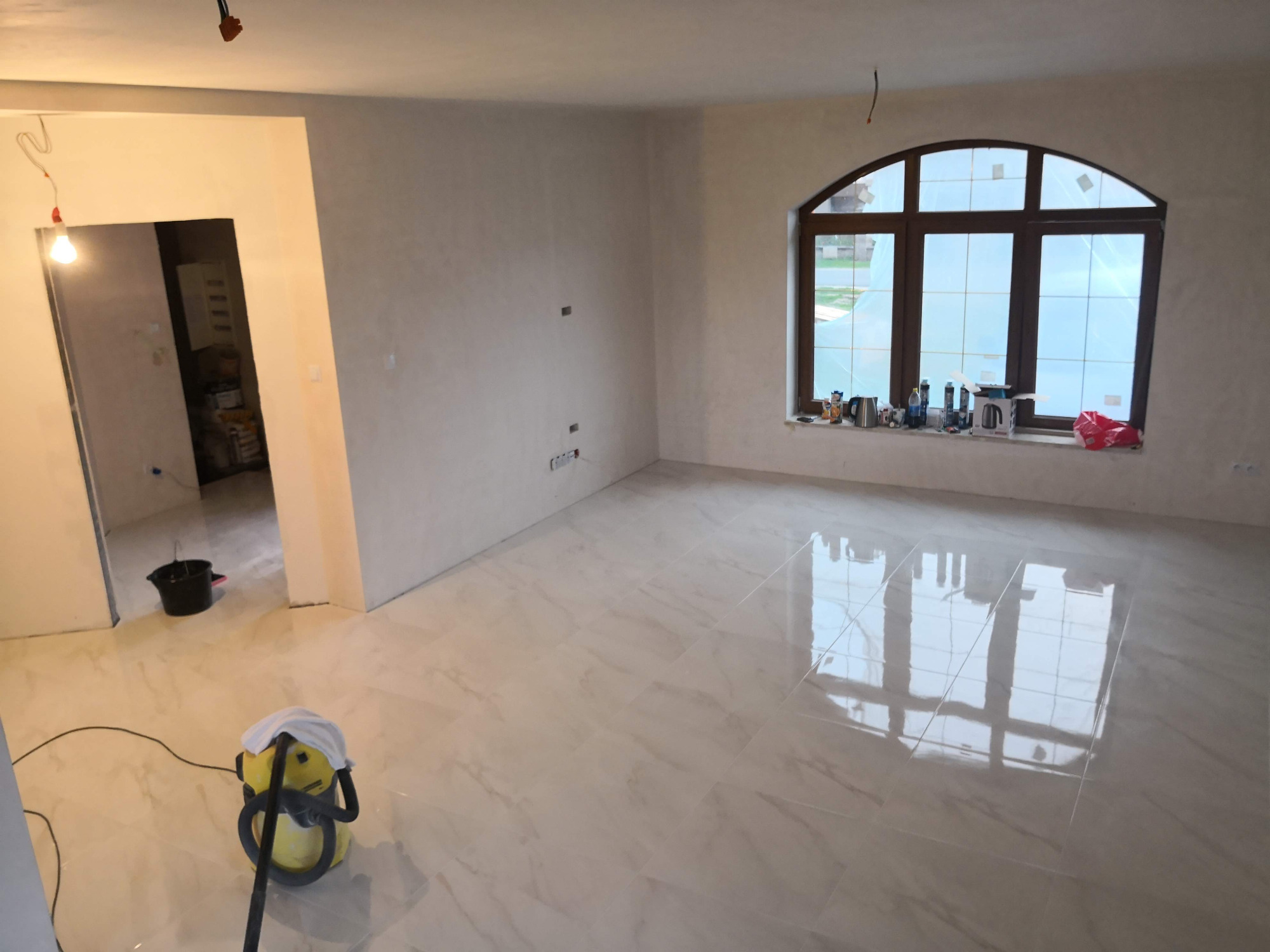 Salon w trakcie prac wykończeniowych: świeżo położona, błyszcząca, jasna podłoga z płytek, odkurzacz przemysłowy na pierwszym planie, okno z drewnianą ramą i parapetem zastawionym przedmiotami w tle.