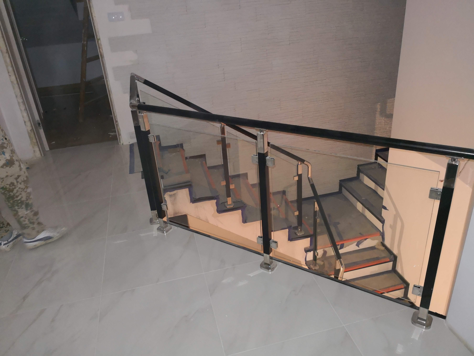 Szklana balustrada schodowa z czarnymi elementami wykończeniowymi, widok z góry na marmurowe płytki podłogowe, częściowo widoczna osoba w odzieży moro.