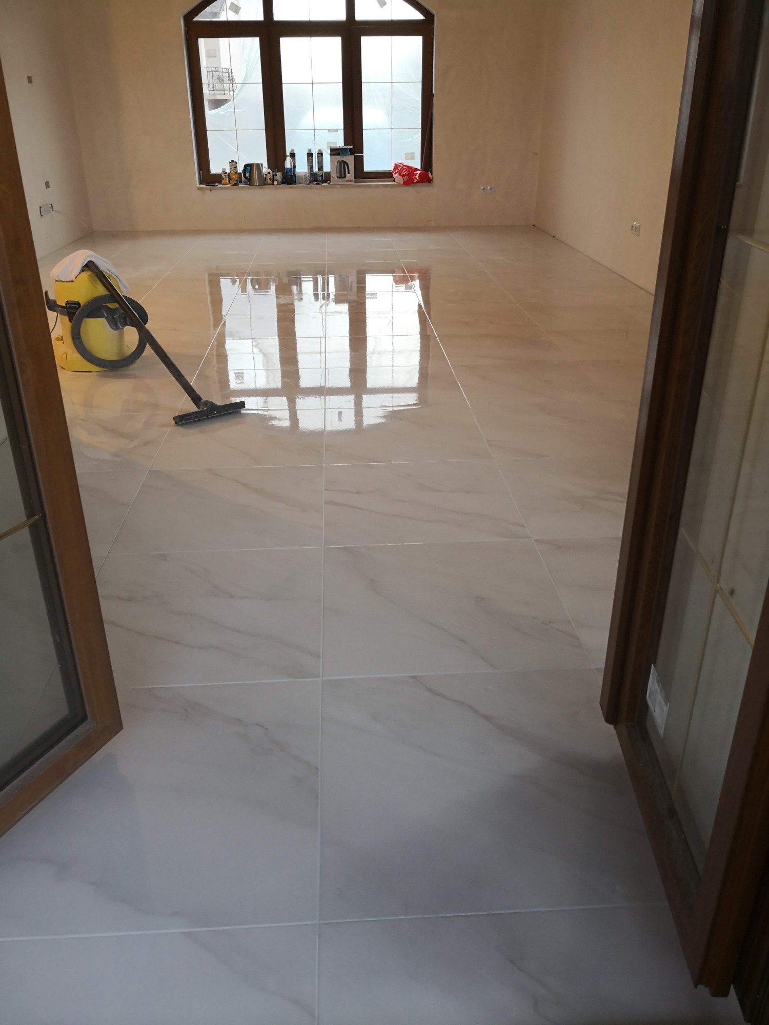 Błyszcząca, beżowo-biała podłoga z płytek imitujących marmur, świeżo po ułożeniu, z widocznym odkurzaczem przemysłowym i odbiciem okna w podłodze.