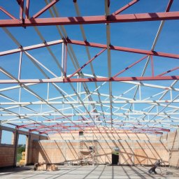WMK-MISTAL Kaczorowski Mirosław - Wnętrze budowanej hali z widoczną konstrukcją stalową dachu w kolorach czerwonym i białym oraz ścianami z cegły dziurawki. Na podłodze widoczne cienie konstrukcji, a w tle błękitne niebo.
