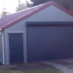 Gotowa hala garażowa z szarej płyty warstwowej z czerwoną blachodachówką, zamykana roletą zewnętrzną i drzwiami wejściowymi, na trawniku z resztkami śniegu.