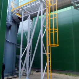Zewnętrzna konstrukcja stalowa z żółtą drabinką, platformą z barierkami i schodami, wspierająca rurociąg na tle zielonej ściany hali.