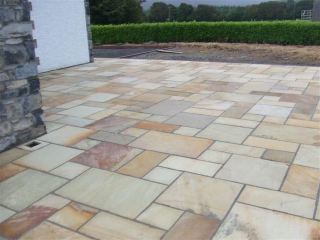 Ułożona z nieregularnych płyt piaskowca nawierzchnia tarasu, widoczna ściana budynku z kamienną okładziną i odpływ liniowy, w tle żywopłot.