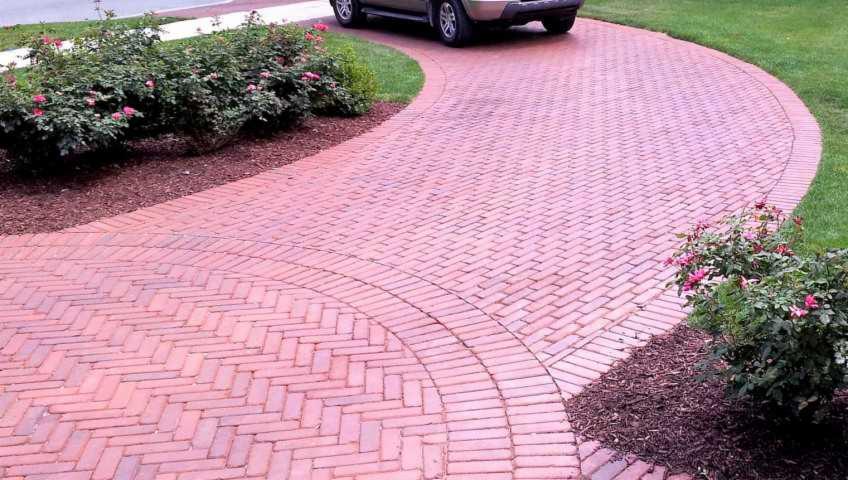 Ceglana, kręta ścieżka z kostki brukowej prowadzi do podjazdu. Widoczne rabaty z krzewami róż i zaparkowany samochód. Kąt ujęcia podkreśla symetrię wzoru.