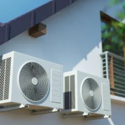Dwa białe klimatyzatory zamontowane na ścianie budynku jednorodzinnego, widoczne z zewnątrz, z fragmentem balkonu i zieleni w tle.