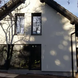 Elewacja domu jednorodzinnego z oknami, w jasnym odcieniu bieli, z widocznymi cieniami drzew. Dach dwuspadowy w kolorze grafitowym. Widoczne płyty na ziemi.