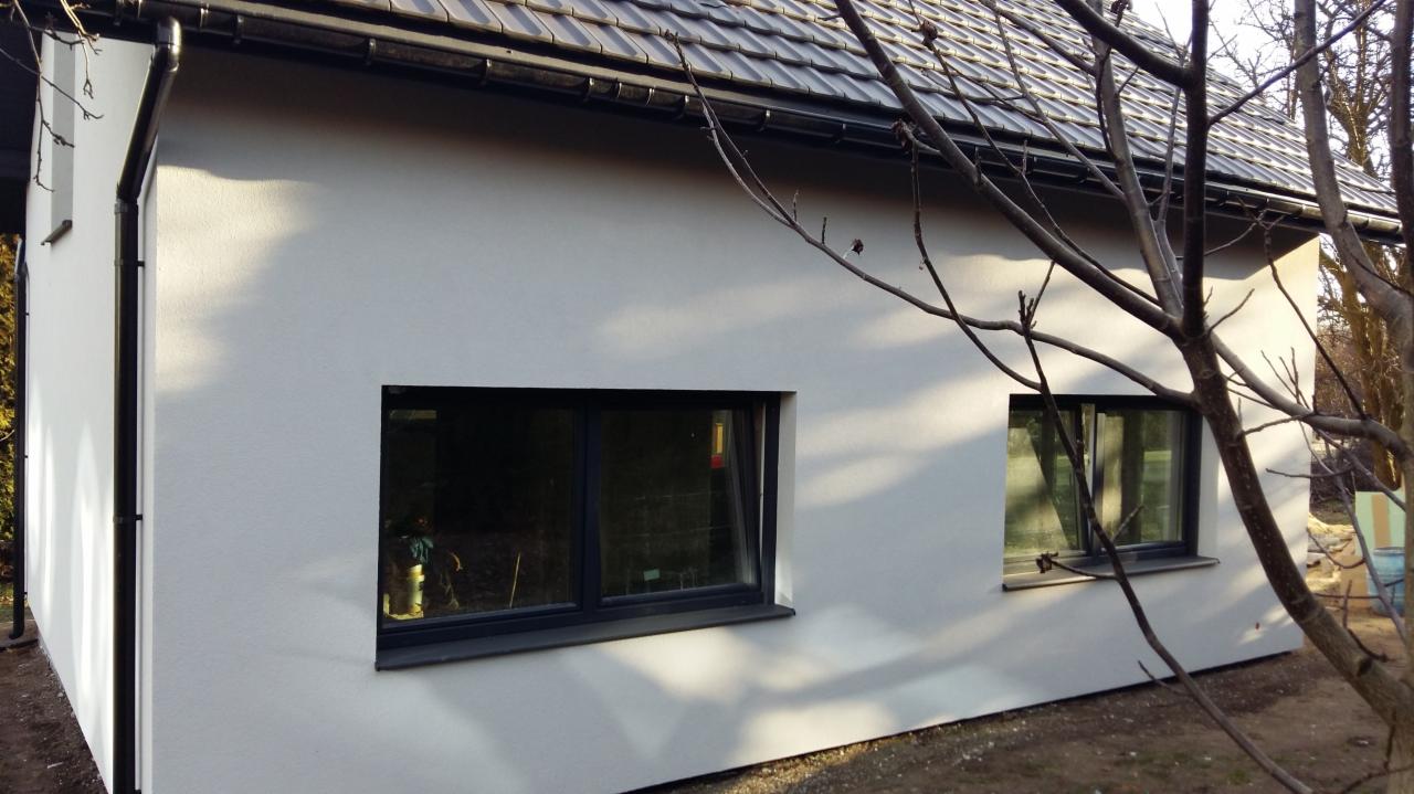 Elewacja budynku po nałożeniu białego tynku, widoczne okna z czarną ramą, rynna i fragment dachu. W tle drzewa bez liści.