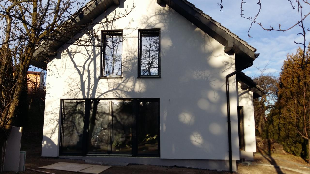 Elewacja domu jednorodzinnego z oknami, w jasnym odcieniu bieli, z widocznymi cieniami drzew. Dach dwuspadowy w kolorze grafitowym. Widoczne płyty na ziemi.