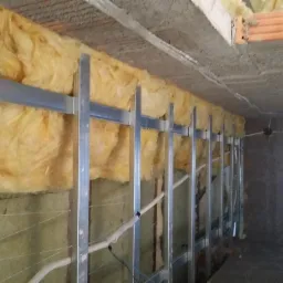 Metalowy stelaż z izolacją z wełny mineralnej na ścianie, przygotowany do montażu płyt gipsowo-kartonowych. Widoczne elementy instalacji elektrycznej.