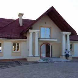Nowo wybudowany, parterowy dom z poddaszem użytkowym, z czerwonym dachem, kolumnami przy wejściu i brukowanym podjazdem. Osoba stoi obok kolumny.