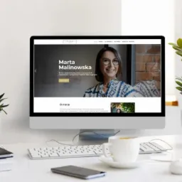 Ekran komputera iMac prezentujący stronę internetową Marty Malinowskiej na białym biurku z roślinami doniczkowymi i filiżanką kawy.