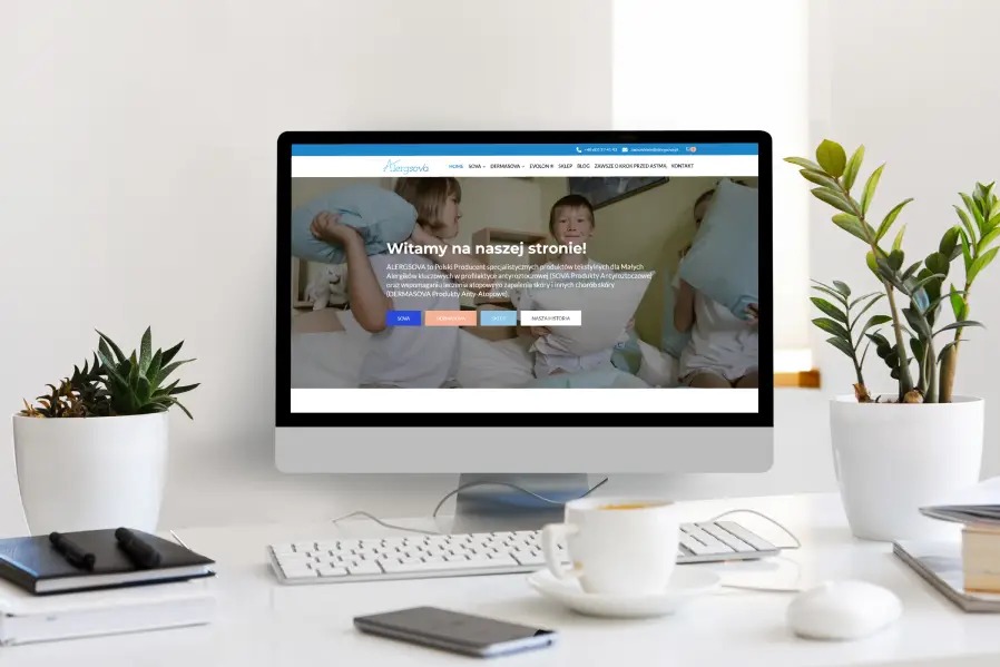 Ekran komputera stacjonarnego prezentuje stronę internetową Alergsova z dziećmi i poduszkami. Obok rośliny doniczkowe, kawa i akcesoria biurowe na białym biurku.