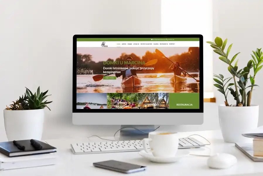 Ekran komputera wyświetla stronę internetową 'Domki u Marcina' z motywem kajaków i domków letniskowych. Na biurku roślina, kawa i akcesoria.
