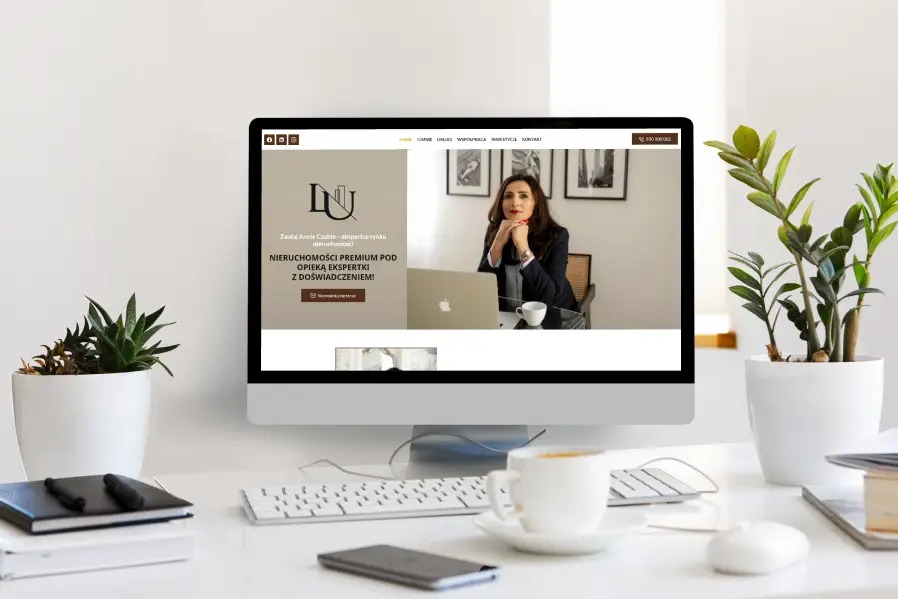 Monitor wyświetla stronę internetową z logo i zdjęciem kobiety, otoczony biurowymi akcesoriami i roślinami doniczkowymi na białym biurku. Minimalistyczny design.