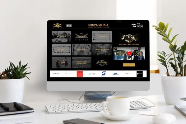 Ekran komputera stacjonarnego prezentuje stronę internetową Grupa Silesia na biurku z kawą i roślinami doniczkowymi. Minimalistyczny design.